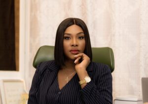 Professional woman in a pinstripe blazer sits at a desk, resting her chin on her hand in a thoughtful pose, wearing a gold cross necklace and watch.