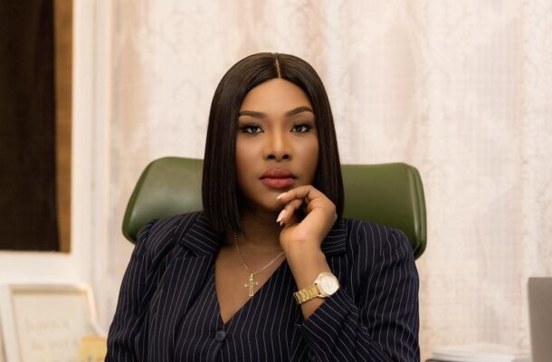 Professional woman in a pinstripe blazer sits at a desk, resting her chin on her hand in a thoughtful pose, wearing a gold cross necklace and watch.