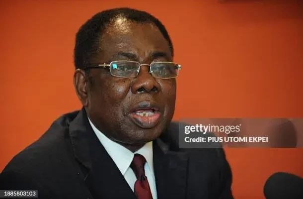 A man in a dark suit and red tie wearing glasses speaks into a microphone against an orange background, as if at a press conference.