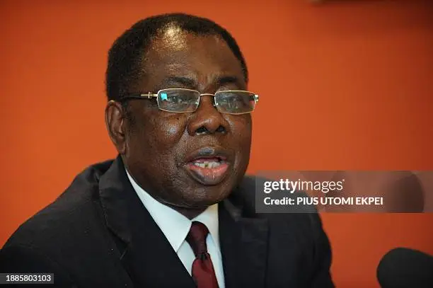 A man in a dark suit and red tie wearing glasses speaks into a microphone against an orange background, as if at a press conference.