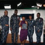 Group photo in a studio: four military personnel in camo uniforms and a woman in a purple dress holding a diploma.