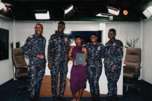 Group photo in a studio: four military personnel in camo uniforms and a woman in a purple dress holding a diploma.