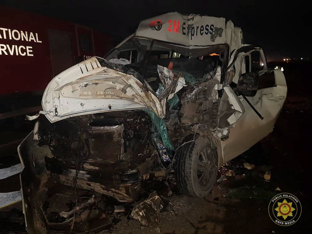 White wrecked van with severe front-end damage sits on a dark highway at night; windshield shattered, exposed engine, door ajar, 2M Express labeled cab ahead of the wreckage.
