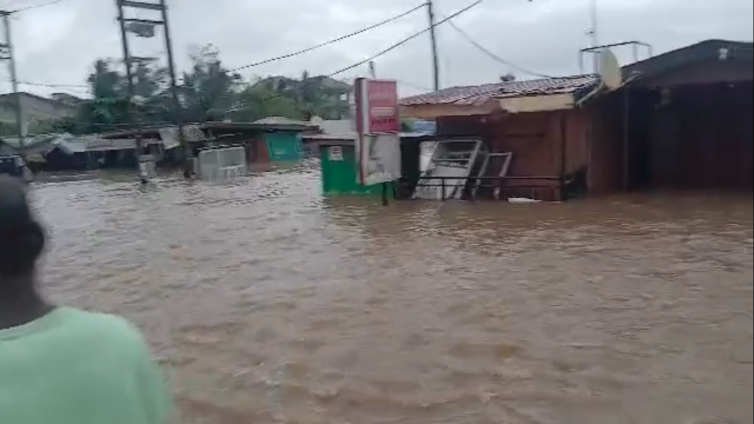 Flooded street in a town with brown water surrounding shopfronts and debris, utility poles, and tangled wires overhead.