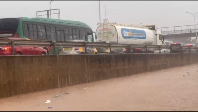 Roadway in heavy rain with traffic jam beside a flooded barrier; a green bus and a white tanker truck are visible.