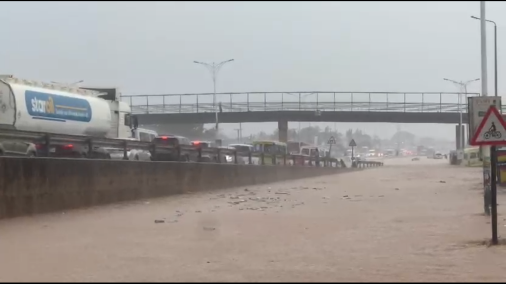 Heavy traffic on a flooded highway with muddy water covering the roadway and an overpass in the distance on a cloudy day.