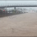 Flooded highway with fast-moving water covering multiple lanes, cars stuck in traffic, an overpass in the background, and a bicycle warning sign on the right.