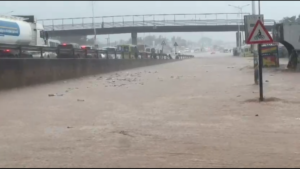 Flooded highway with fast-moving water covering multiple lanes, cars stuck in traffic, an overpass in the background, and a bicycle warning sign on the right.