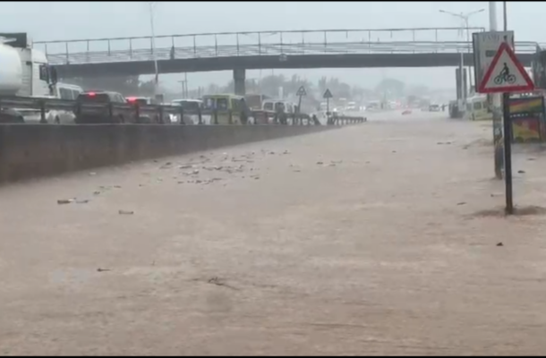 Flooded highway with fast-moving water covering multiple lanes, cars stuck in traffic, an overpass in the background, and a bicycle warning sign on the right.