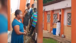 People line up along a narrow orange-walled outdoor corridor; a woman in a blue dress stands in the foreground while others blur in the background.