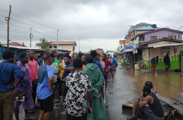 Group of people standing in a flooded street of a neighborhood, watching and talking amid muddy water and colorful buildings.