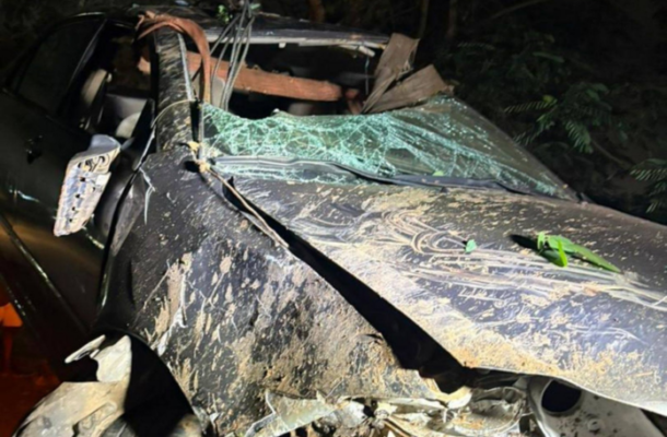 Nighttime scene of a black SUV with severe front-end damage and a shattered windshield, attached to a tow crane in a muddy, wooded roadside area.
