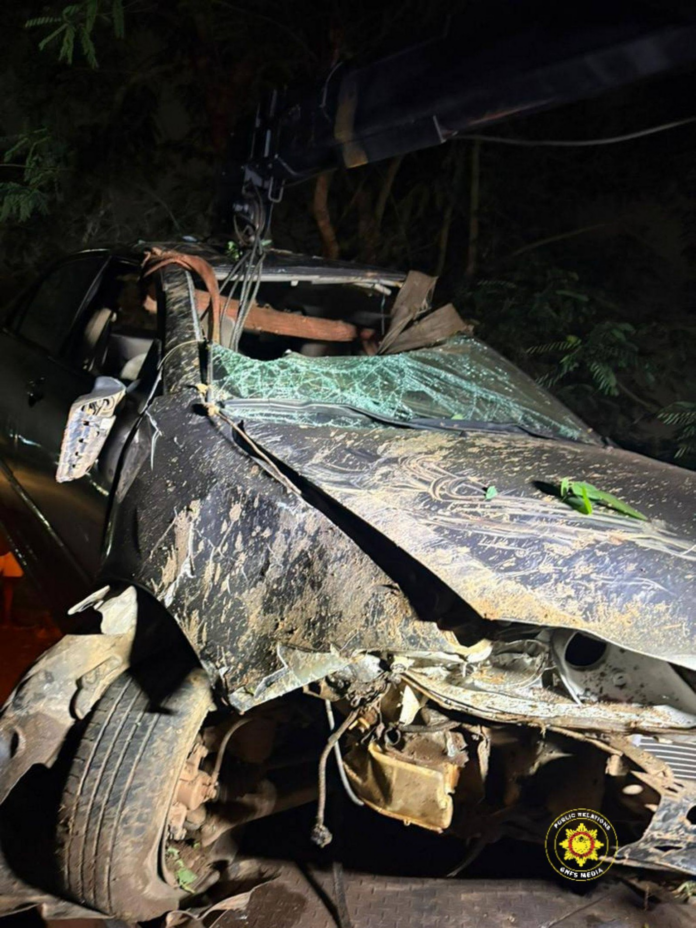Nighttime scene of a black SUV with severe front-end damage and a shattered windshield, attached to a tow crane in a muddy, wooded roadside area.