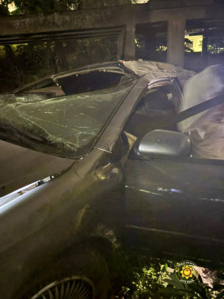 SUV with a shattered windshield and crumpled roof after a crash, driver-side door open, nighttime under an overpass/bridge area .