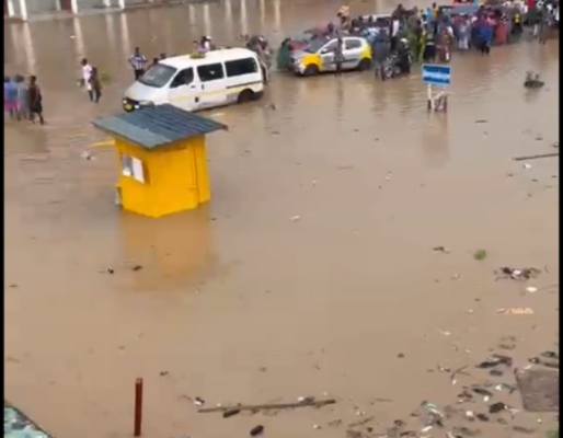 image-2026-06-10T014715378 | Ghanaguardian.com Flooded street with knee-deep water, people gathered along the road as stranded vehicles sit in floodwater, a bright yellow kiosk in the foreground and a building under construction in the background.