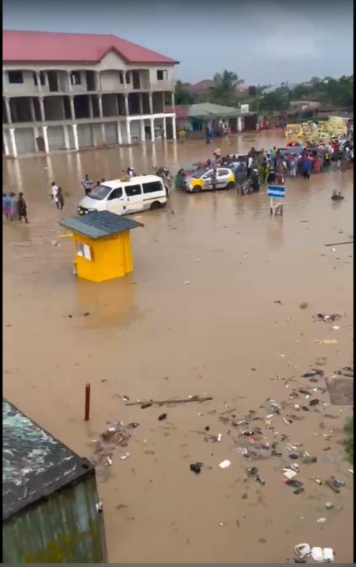 Flooded street with knee-deep water, people gathered along the road as stranded vehicles sit in floodwater, a bright yellow kiosk in the foreground and a building under construction in the background.
