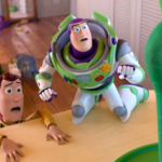 Buzz Lightyear and Woody lean over a table edge with surprised expressions as a tall green toy stands nearby; a cluttered toy room is in the background.