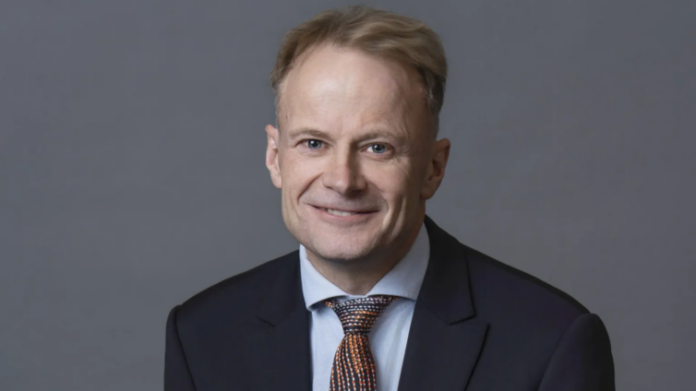 Portrait of a smiling man in a dark suit and patterned tie against a gray background.