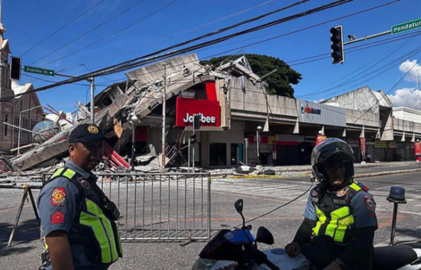 Two police officers in high-visibility vests stand behind barricades on a street in front of a collapsed shopping center with a red Jollibee sign, blue sky above.