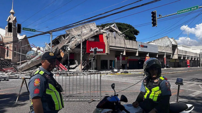 Two police officers in high-visibility vests stand behind barricades on a street in front of a collapsed shopping center with a red Jollibee sign, blue sky above.