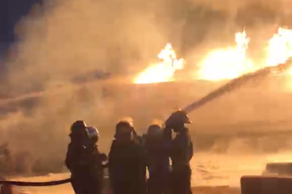 image-327 | Ghanaguardian.com Firefighters in turnout gear directing a hose at a large blaze at night, with bright flames and thick smoke behind them.