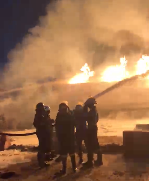 Firefighters in turnout gear directing a hose at a large blaze at night, with bright flames and thick smoke behind them.
