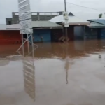 Flooded street in a small commercial area with water covering the road and storefronts, reflecting signs and buildings on the surface.