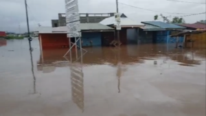 Flooded street in a small commercial area with water covering the road and storefronts, reflecting signs and buildings on the surface.