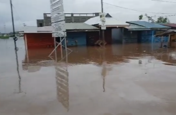 Flooded street in a small commercial area with water covering the road and storefronts, reflecting signs and buildings on the surface.
