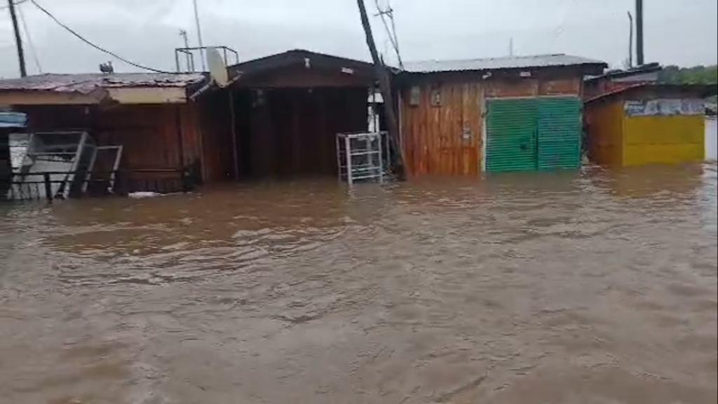 Row of small wooden storefronts partially submerged in brown floodwater with overhanging roofs and scattered debris nearby.
