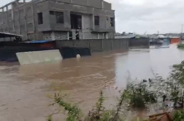 Flooded street scene with muddy water surrounding a partially built concrete building and nearby barriers.