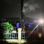 Nighttime scene of a fenced area with barbed wire, a tall pole emitting smoke, and a distant structure lit in blue lights; an emergency badge is in the lower right.