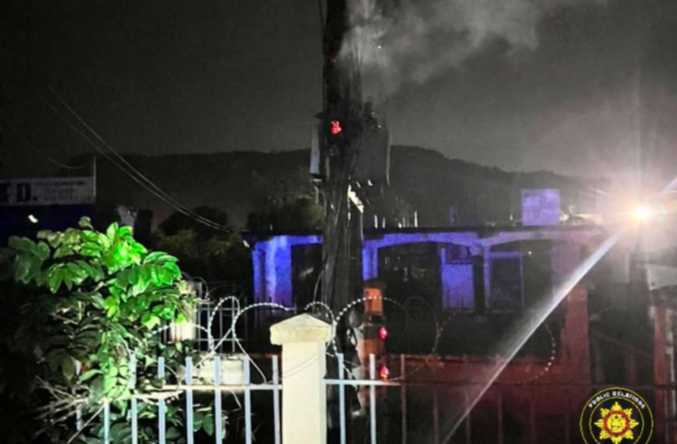 Nighttime scene of a fenced area with barbed wire, a tall pole emitting smoke, and a distant structure lit in blue lights; an emergency badge is in the lower right.
