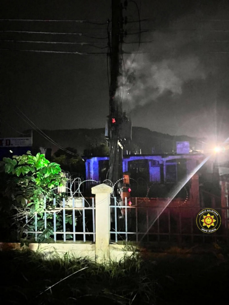 Nighttime scene of a fenced area with barbed wire, a tall pole emitting smoke, and a distant structure lit in blue lights; an emergency badge is in the lower right.