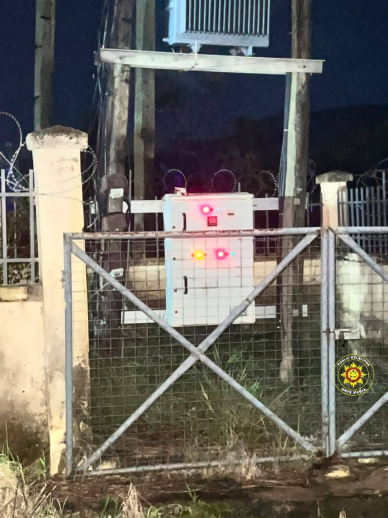 Security gate and wire fence surround an electrical control cabinet with glowing red and yellow indicator lights at night.