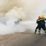 Firefighters in protective gear spraying water to control a blaze on a smoky road scene.