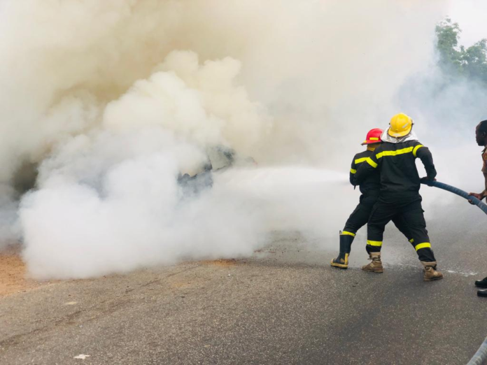 Firefighters in protective gear spraying water to control a blaze on a smoky road scene.