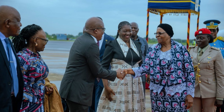 Group of formally dressed individuals on an airport tarmac shake hands, with a woman in a patterned dress smiling nearby and others watching the greeting under a blue-and-gold ceremonial canopy.