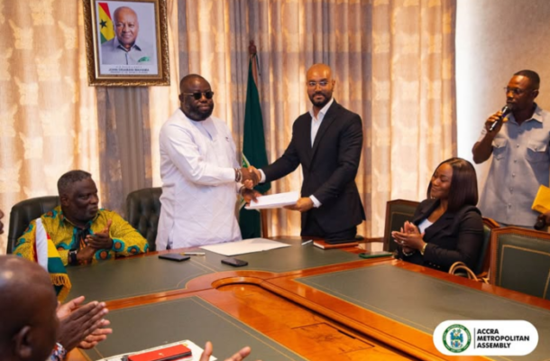 Two men shake hands across a conference table after exchanging documents in a formal signing ceremony, with the Accra Metropolitan Assembly logo visible in the corner and others seated applauding.