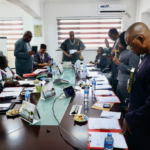 Group of people gathered around a long conference table, some standing with hands raised in prayer or blessing during a meeting.