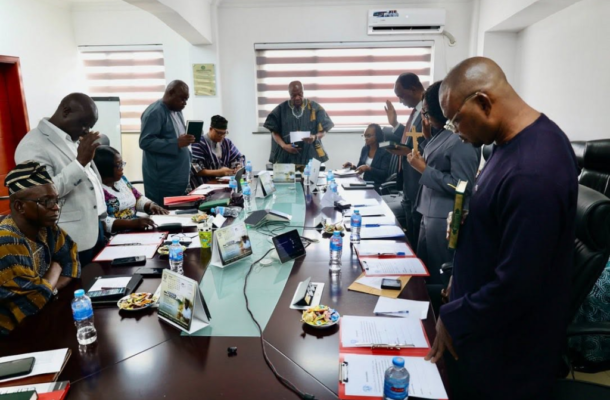 Group of people gathered around a long conference table, some standing with hands raised in prayer or blessing during a meeting.