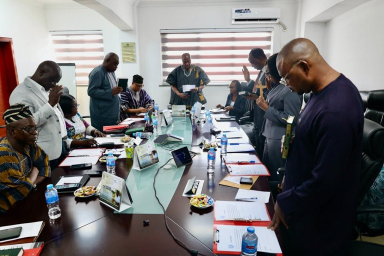 Group of people gathered around a long conference table, some standing with hands raised in prayer or blessing during a meeting.