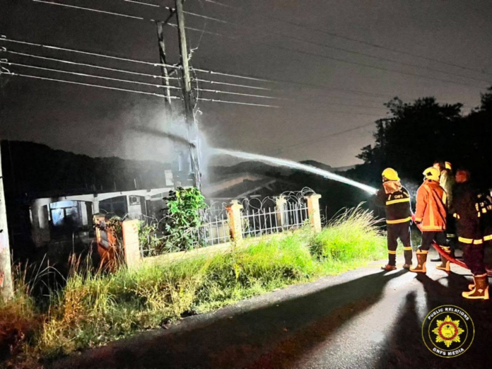 Nighttime firefighting scene: firefighters spray water from a hose toward a burning or smoldering building behind a fence and power lines.