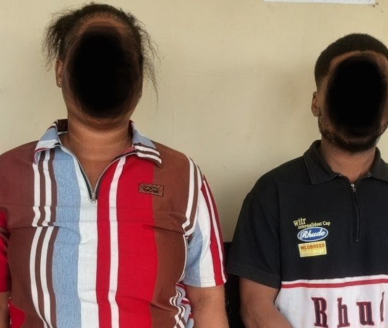 Two people stand side by side against a beige wall; their faces are obscured. The left person wears a red, white, blue striped polo, the right person wears a dark polo with patches.
