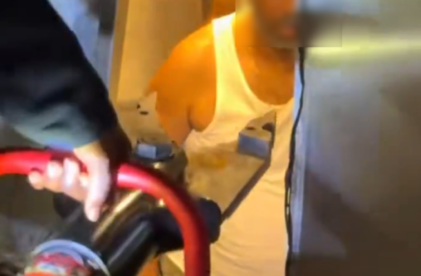 Unactioned scene: a person in a white tank top and light shorts stands in a doorway, facing the camera, with a red-handled cart or buggy in the foreground and another figure partially visible on the left. Face intentionally blurred for privacy.