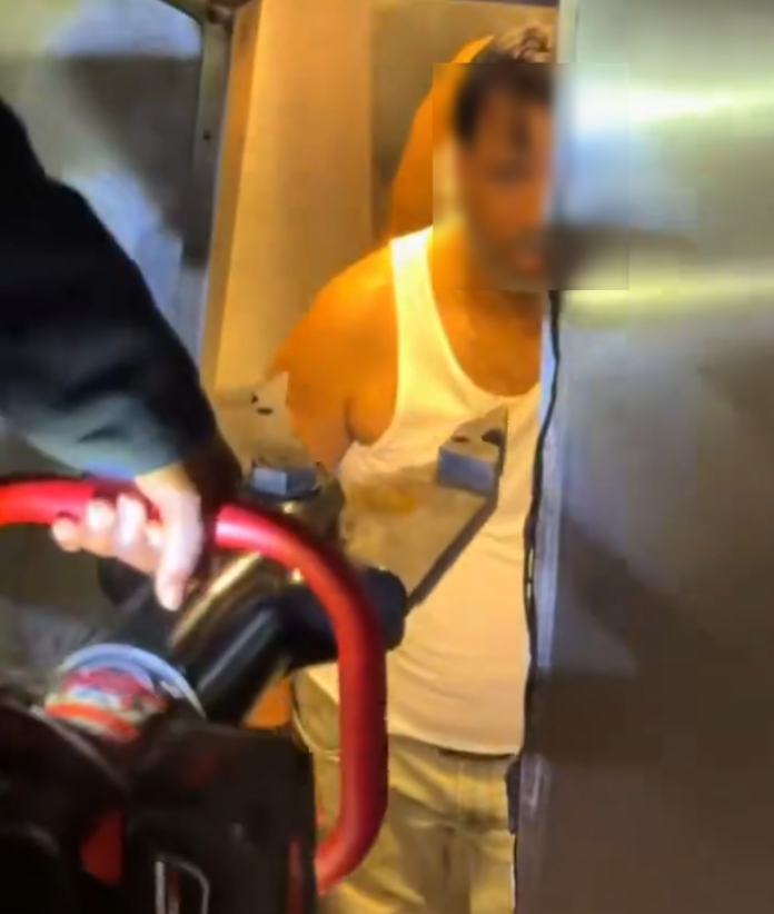 Unactioned scene: a person in a white tank top and light shorts stands in a doorway, facing the camera, with a red-handled cart or buggy in the foreground and another figure partially visible on the left. Face intentionally blurred for privacy.