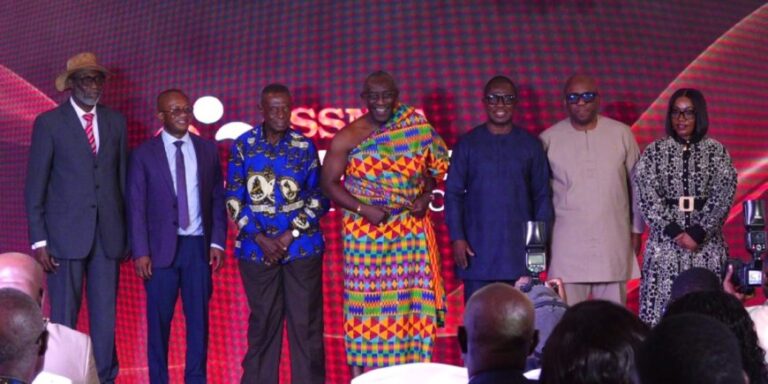 Seven people pose on a stage with a red patterned backdrop during a formal event.