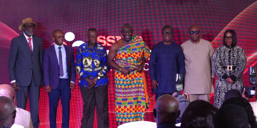 Seven people pose on a stage with a red patterned backdrop during a formal event.