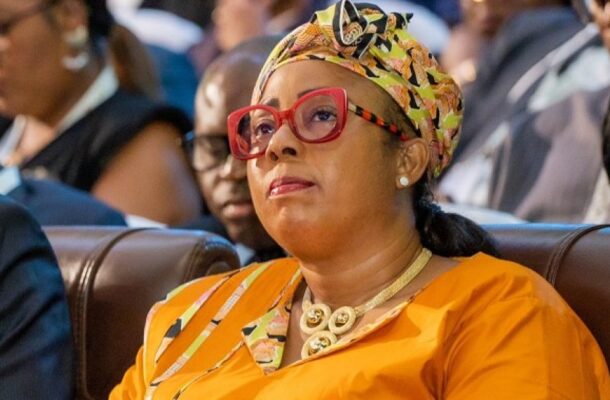 image_870x_6a2e9bdc499d1 | Ghanaguardian.com Woman in orange attire with a patterned headwrap and red glasses sitting in an audience, looking attentive.