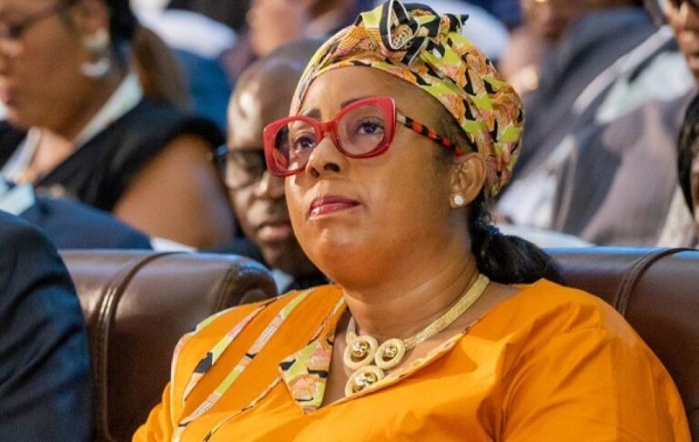 Woman in orange attire with a patterned headwrap and red glasses sitting in an audience, looking attentive.